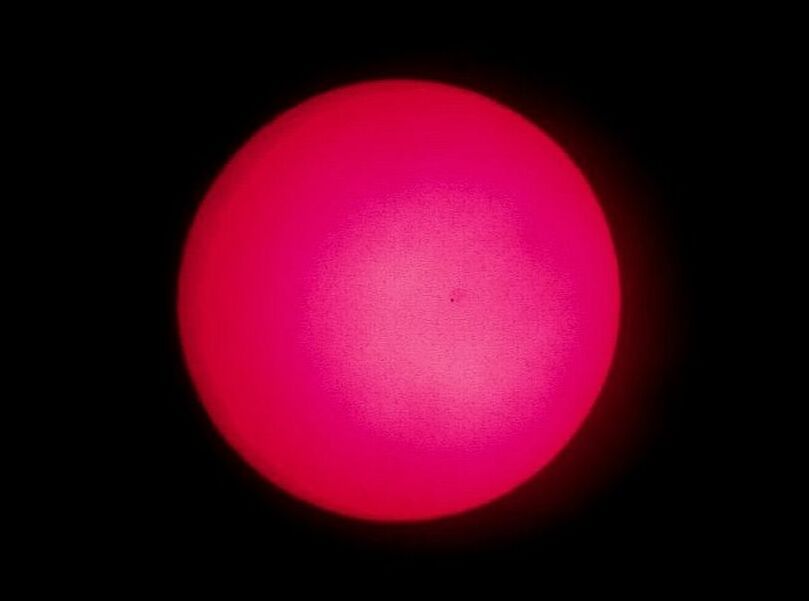 Fotografía del tránsito de Mercurio a través de un telescopio solar. El Sol es una gran bola roja sobre un fondo negro y Mercurio un minúsculo puntito negro un poco a la derecha del centro.
